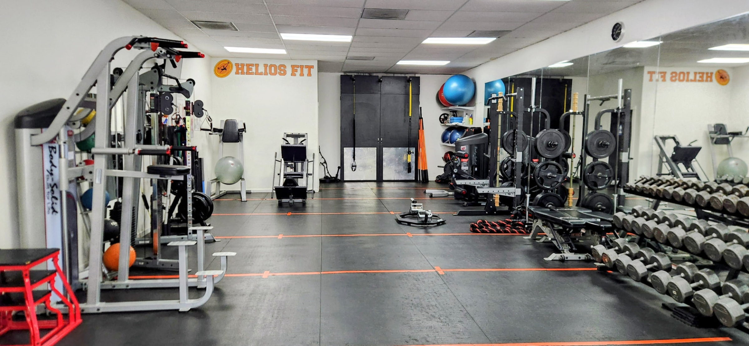 Helios Fit logo on the studio wall with racks, dumbbells and turf lanes